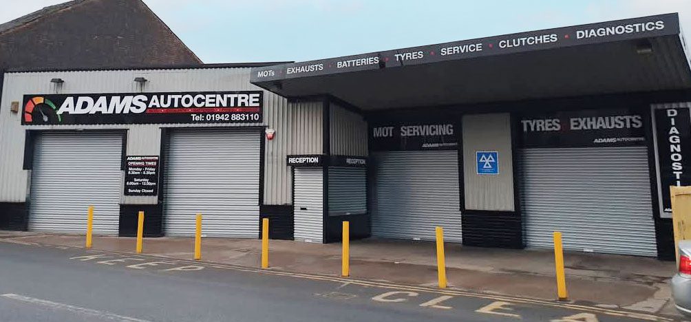 Adams Autocentre garage front. There are yellow parking bollards in front, and the garage has 4 large doors for vehicles to use.